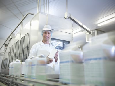 Dans un environnement industriel propre et éclairé, une personne vêtue d’une blouse blanche, d’une charlotte et d’un couvre-chef tient une brique de produit devant une machine de conditionnement. Au premier plan, plusieurs emballages apparaissent flous en mouvement sur un convoyeur, illustrant une chaîne de production automatisée.
