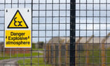 Panneau d’avertissement jaune indiquant « Danger explosive atmosphere » et symbole de risque d’explosion, fixé sur une grille métallique. À l’arrière-plan, une zone clôturée et des bâtiments industriels sont visibles mais flous.