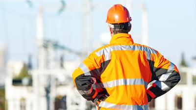 Personne vue de dos portant un casque de sécurité orange et une veste haute visibilité orange avec bandes réfléchissantes, debout devant des installations industrielles floues, probablement sur un site électrique ou énergétique.