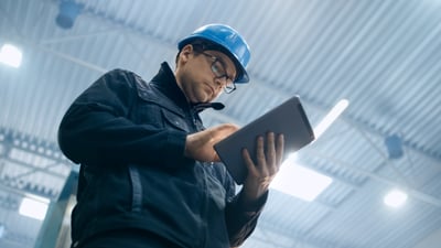 Vue en contre-plongée d’une personne équipée d’un casque de sécurité bleu, de lunettes et d’une veste de travail sombre, consultant une tablette numérique à l’intérieur d’un site industriel. Le plafond métallique et les luminaires visibles en arrière-plan suggèrent un atelier ou un entrepôt.