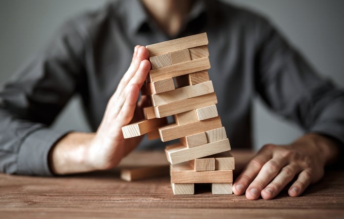 Vue rapprochée d’une personne assise derrière une table en bois, tenant la main près d’une tour de blocs de bois empilés de manière instable. La structure penche sur le côté tandis que la main semble la soutenir pour éviter qu’elle ne s’écroule. L’image évoque la maîtrise des risques, la résilience ou le maintien de l’équilibre face à une situation fragile.