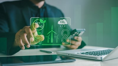 Personne utilisant un smartphone devant un ordinateur portable, avec une interface graphique verte superposée illustrant des thèmes de développement durable : « Net Zero », réduction du CO₂, éoliennes, planète tenue dans des mains et économie circulaire. L’image évoque le suivi ou la gestion numérique d’objectifs environnementaux.