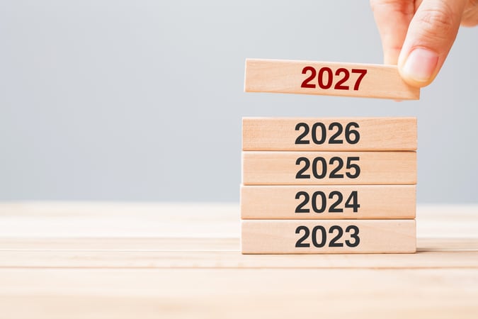 Une main place un bloc en bois marqué « 2027 » au sommet d’une pile de blocs portant les années 2023, 2024, 2025 et 2026, illustrant le passage à une nouvelle année, la planification ou la projection vers l’avenir.