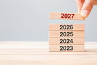 Une main place un bloc en bois marqué « 2027 » au sommet d’une pile de blocs portant les années 2023, 2024, 2025 et 2026, illustrant le passage à une nouvelle année, la planification ou la projection vers l’avenir.