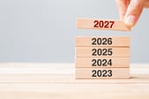 Une main place un bloc en bois marqué « 2027 » au sommet d’une pile de blocs portant les années 2023, 2024, 2025 et 2026, illustrant le passage à une nouvelle année, la planification ou la projection vers l’avenir.
