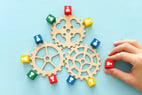 Trois engrenages en bois entourés de cubes colorés représentant des personnes, avec une main plaçant un cube rouge près du mécanisme.
