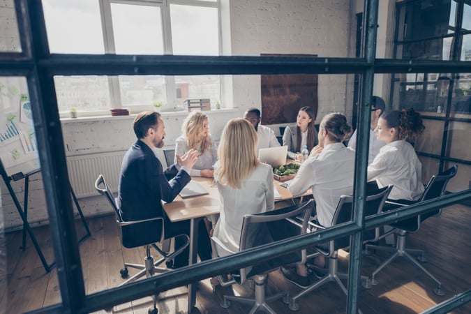 Groupe de personnes réunies autour d’une table dans une salle de réunion vitrée, avec des ordinateurs portables et un tableau présentant des graphiques.