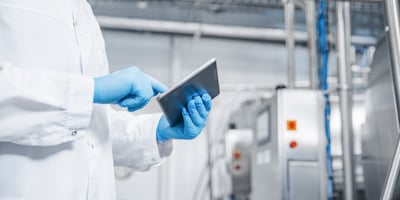 Gros plan sur une personne vêtue d’une blouse blanche et de gants de protection bleus, tenant une tablette tactile et interagissant avec son écran. L’image est prise dans un environnement industriel propre, avec des équipements en acier inoxydable, des câbles et des panneaux de commande visibles en arrière-plan. L’ensemble évoque des activités de contrôle, de suivi ou de gestion de processus de production.
