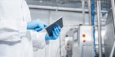 Gros plan sur une personne vêtue d’une blouse blanche et de gants de protection bleus, tenant une tablette tactile et interagissant avec son écran. L’image est prise dans un environnement industriel propre, avec des équipements en acier inoxydable, des câbles et des panneaux de commande visibles en arrière-plan. L’ensemble évoque des activités de contrôle, de suivi ou de gestion de processus de production.