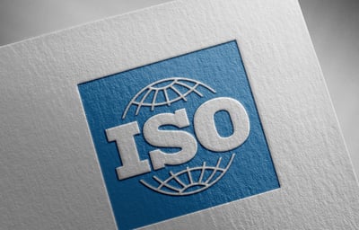 Vue en perspective d’un logo ISO en relief blanc placé sur un carré bleu. Un globe stylisé entoure partiellement les lettres « ISO ». L’ensemble est imprimé ou embossé sur un support blanc à texture papier, photographié en gros plan.