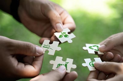 Gros plan sur plusieurs mains tenant et assemblant des pièces de puzzle ornées d’icônes environnementales, dont le recyclage, une éolienne, une prise électrique avec des feuilles et une usine verte. L’image symbolise la coopération et les initiatives en matière de durabilité et de protection de l’environnement.