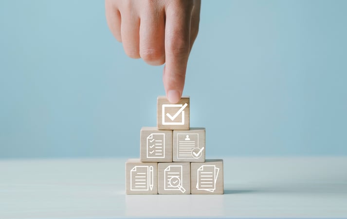Main appuyant sur un cube en bois marqué d’une coche, au sommet d’un empilement de cubes illustrant des documents, listes de contrôle et validations. 