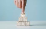 Main appuyant sur un cube en bois marqué d’une coche, au sommet d’un empilement de cubes illustrant des documents, listes de contrôle et validations. 