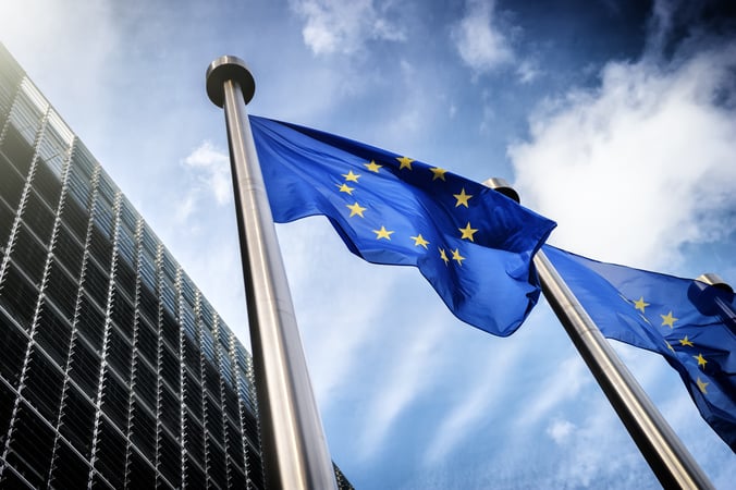 Drapeaux de l’Union européenne flottant au vent devant un bâtiment moderne, sous un ciel partiellement nuageux.