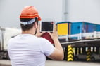 Personne portant un casque de sécurité orange et tenant une tablette numérique, vue de dos, devant une zone de chargement avec des conteneurs de transport en arrière-plan. L’image illustre l’utilisation d’outils numériques dans un environnement logistique ou industriel.