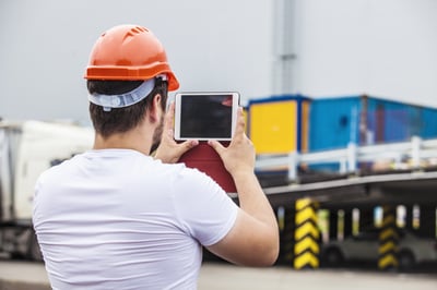 Personne portant un casque de sécurité orange et tenant une tablette numérique, vue de dos, devant une zone de chargement avec des conteneurs de transport en arrière-plan. L’image illustre l’utilisation d’outils numériques dans un environnement logistique ou industriel.
