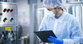 Dans un atelier industriel propre, une personne vêtue d’une combinaison de protection, d’une charlotte, d’un masque et de gants bleus consulte une tablette. À proximité se trouvent un panneau de commande et un équipement de production en acier inoxydable, suggérant une opération de contrôle ou de suivi qualité.