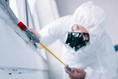 Personne vêtue d'une combinaison de protection blanche et portant un masque respiratoire, effectuant un travail de nettoyage ou de prélèvement à l'aide d'un outil sur une surface blanche.