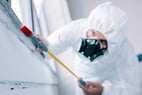 Personne vêtue d'une combinaison de protection blanche et portant un masque respiratoire, effectuant un travail de nettoyage ou de prélèvement à l'aide d'un outil sur une surface blanche.