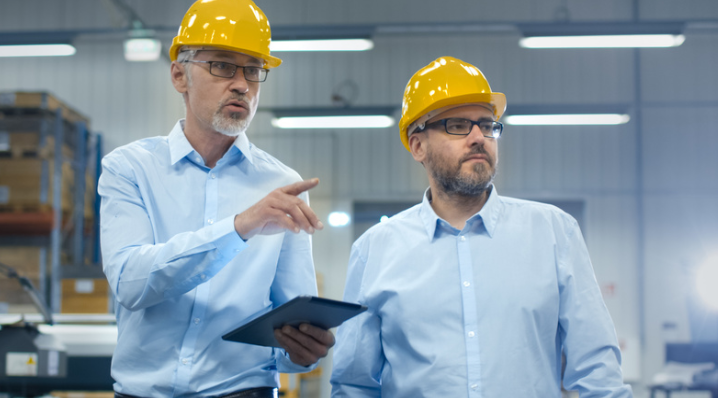 Deux personnes portant des casques de sécurité jaunes et des lunettes de protection se tiennent dans un atelier industriel. L’une d’elles consulte une tablette et désigne une zone de l’usine, tandis que l’autre observe dans la même direction. Des machines et des rayonnages sont visibles à l’arrière-plan.