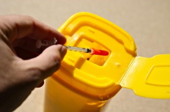 Une main jetant une seringue usagée dans un conteneur jaune destiné aux déchets médicaux à risque biologique. Le couvercle du conteneur est ouvert et conçu pour permettre l’élimination sécurisée des objets tranchants.